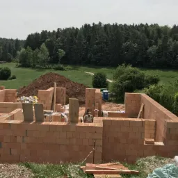 Budowa domu jednorodzinnego z czerwonej cegły, widoczne ściany fundamentowe, w tle zalesiony teren i przenośna toaleta.