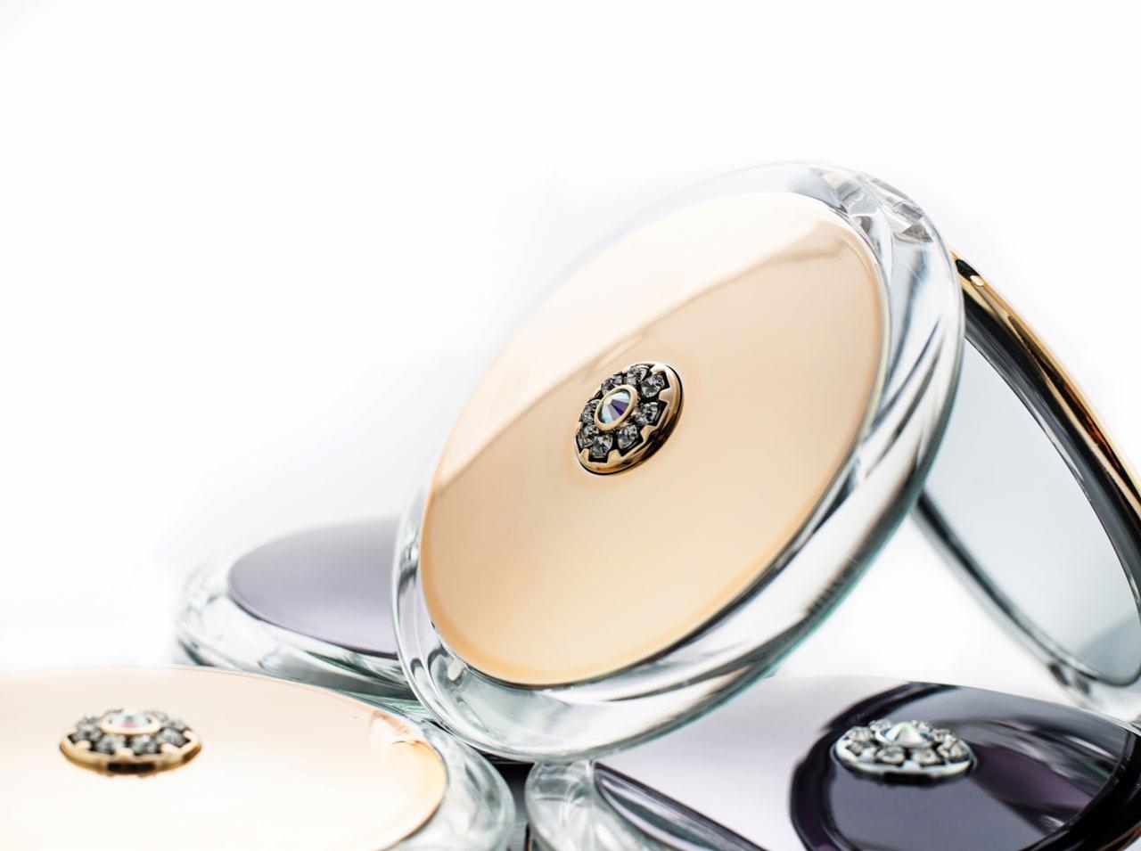 Eleganckie, okrągłe puderniczki z przezroczystego akrylu, zdobione kwiatowym motywem z cyrkoniami na złotym tle, w różnych odcieniach, ułożone na białym tle.