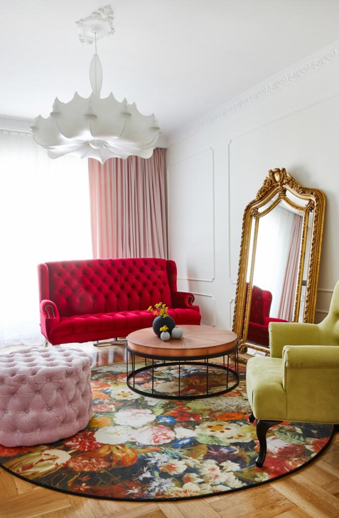 Elegancki salon z czerwonym welurowym fotelem Chesterfield, okrągłym dywanem w kwiaty, zielonym fotelem i lustrem w złotej ramie, oświetlony dekoracyjną lampą.