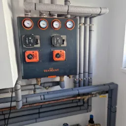 Szara skrzynka sterownicza Termojet z manometrami i zaworami, otoczona zaizolowanymi rurami instalacji grzewczej i hydraulicznej, zamontowana na białej ścianie.