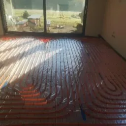 Pomarańczowe rury ogrzewania podłogowego ułożone na siatce, pokryte folią, widok na ogród przez duże okno.