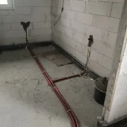 Czerwone rury instalacji hydraulicznej prowadzone po szarym betonowym podłożu w surowym wnętrzu z białych bloczków, widoczne otwory w ścianach z pianką montażową, czarne wykończenie podłogi przy...