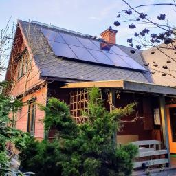 ETER ENERGIA Sp. z o.o. - Dom z panelami słonecznymi na dachu, widok z poziomu ogrodu, z drzewami iglastymi w pierwszym planie i kominem z czerwonej cegły.