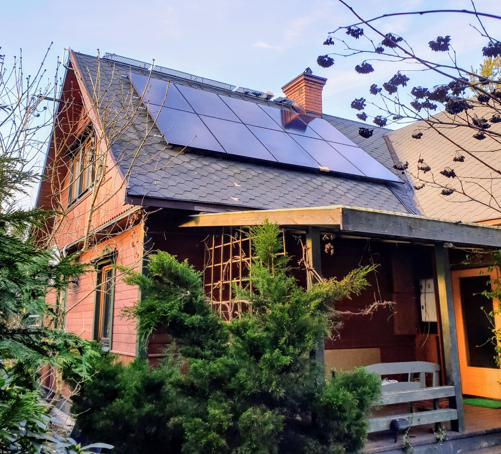 Dom z panelami słonecznymi na dachu, widok z poziomu ogrodu, z drzewami iglastymi w pierwszym planie i kominem z czerwonej cegły.