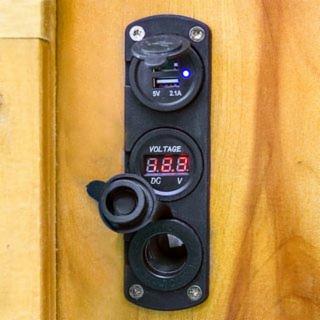 Panel z gniazdem USB, wskaźnikiem napięcia i gniazdem zapalniczki samochodowej, zamontowany na drewnianej powierzchni. Widoczne detale: niebieska dioda LED w gnieździe USB, wyświetlacz wskaźnika...