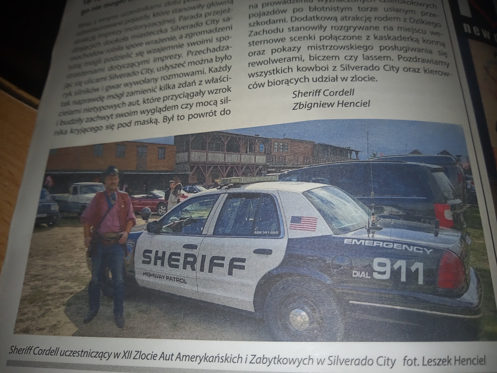 Kowboj w stroju szeryfa stoi obok radiowozu Highway Patrol na Zlocie Aut Amerykańskich i Zabytkowych w Silverado City, widoczny numer 911 i flaga USA na samochodzie.