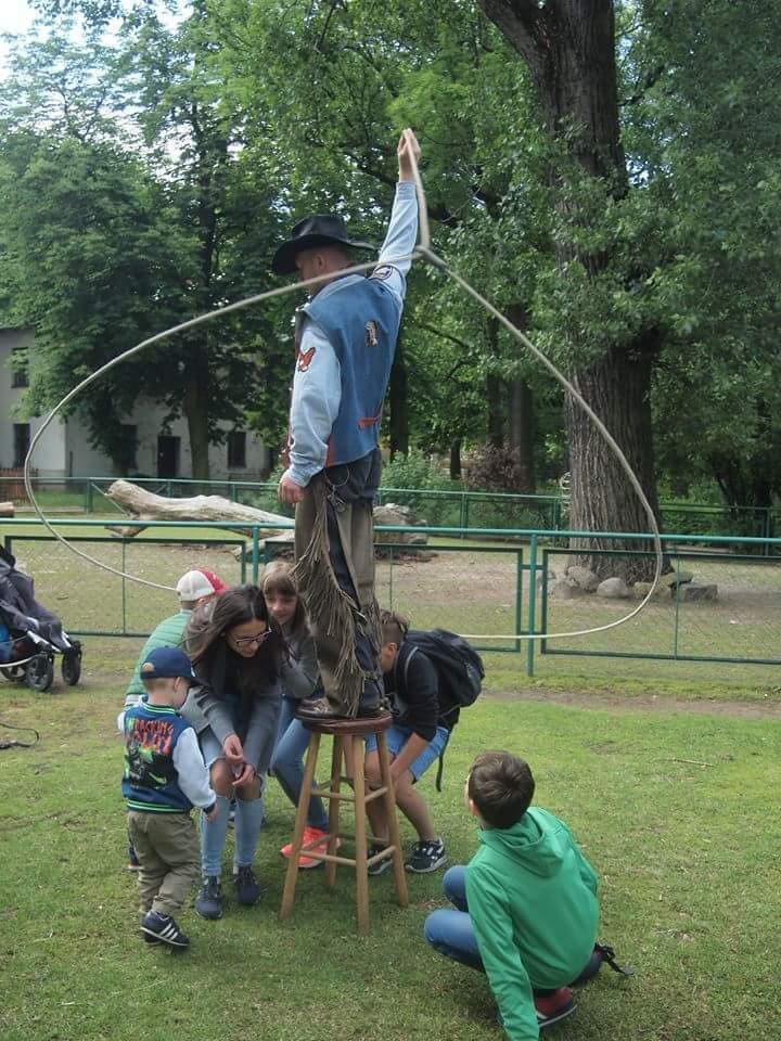 Animator w stroju kowbojskim stoi na stołku i kręci lassem, otoczony grupą dzieci na trawie w parku.