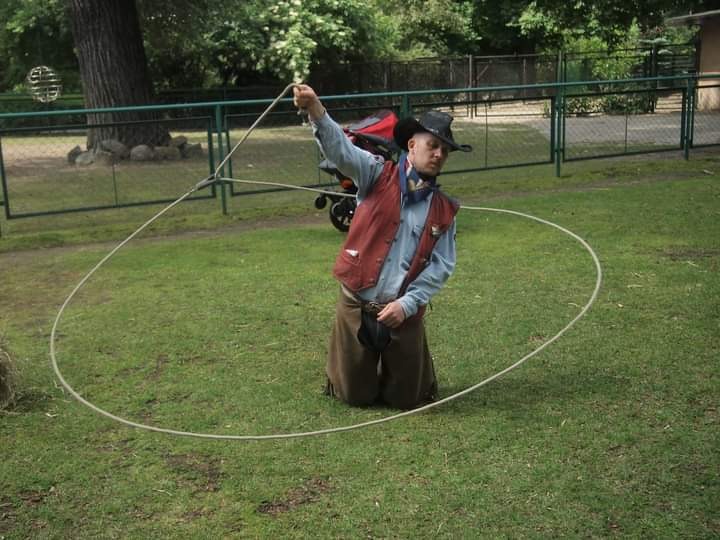 Mężczyzna w stroju kowboja, klęczący na trawie, kręci lassem podczas występu plenerowego.