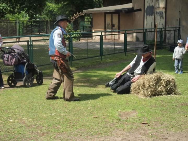 Scena z westernu: mężczyzna w stroju kowboja celuje z rewolweru w leżącego na sianie mężczyznę, w tle wózek dziecięcy i dziecko.