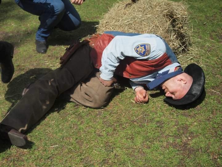 Mężczyzna w stroju kowboja leży na trawie obok stogu siana, udając nieżywego podczas zabawy.