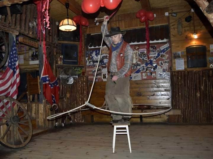 Mężczyzna w stroju kowboja na krześle, wykonujący triki z lassem w rustykalnym wnętrzu z dekoracjami westernowymi, w tym flagami i balonami.