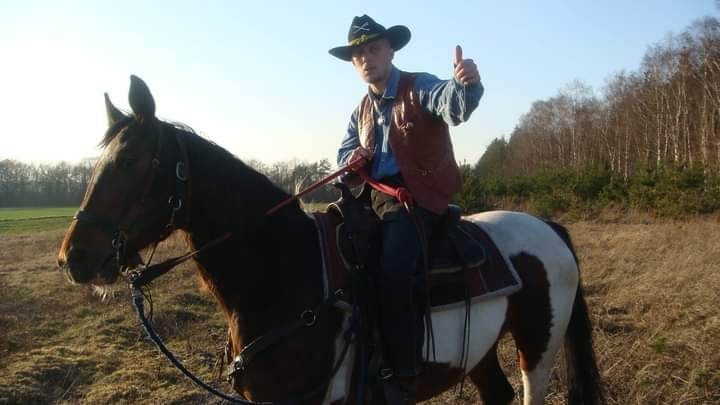 Mężczyzna w kapeluszu kowbojskim i skórzanej kamizelce siedzi na koniu łaciatej maści, pokazując kciuk w górę na tle łąki z drzewami w oddali.