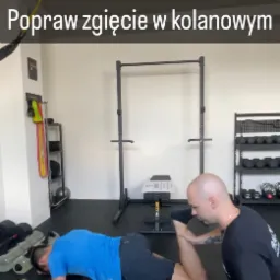 Mężczyzna leżący na brzuchu na macie, używający rollera do rozluźniania mięśni czworogłowych uda, podczas gdy trener personalny delikatnie podnosi jego nogę, wspomagając rozciąganie w siłowni.