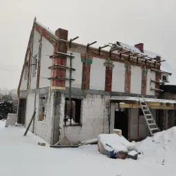Budowa domu jednorodzinnego w stanie surowym zimą, widoczne białe bloczki, drewniane wzmocnienia, rusztowania i śnieg.