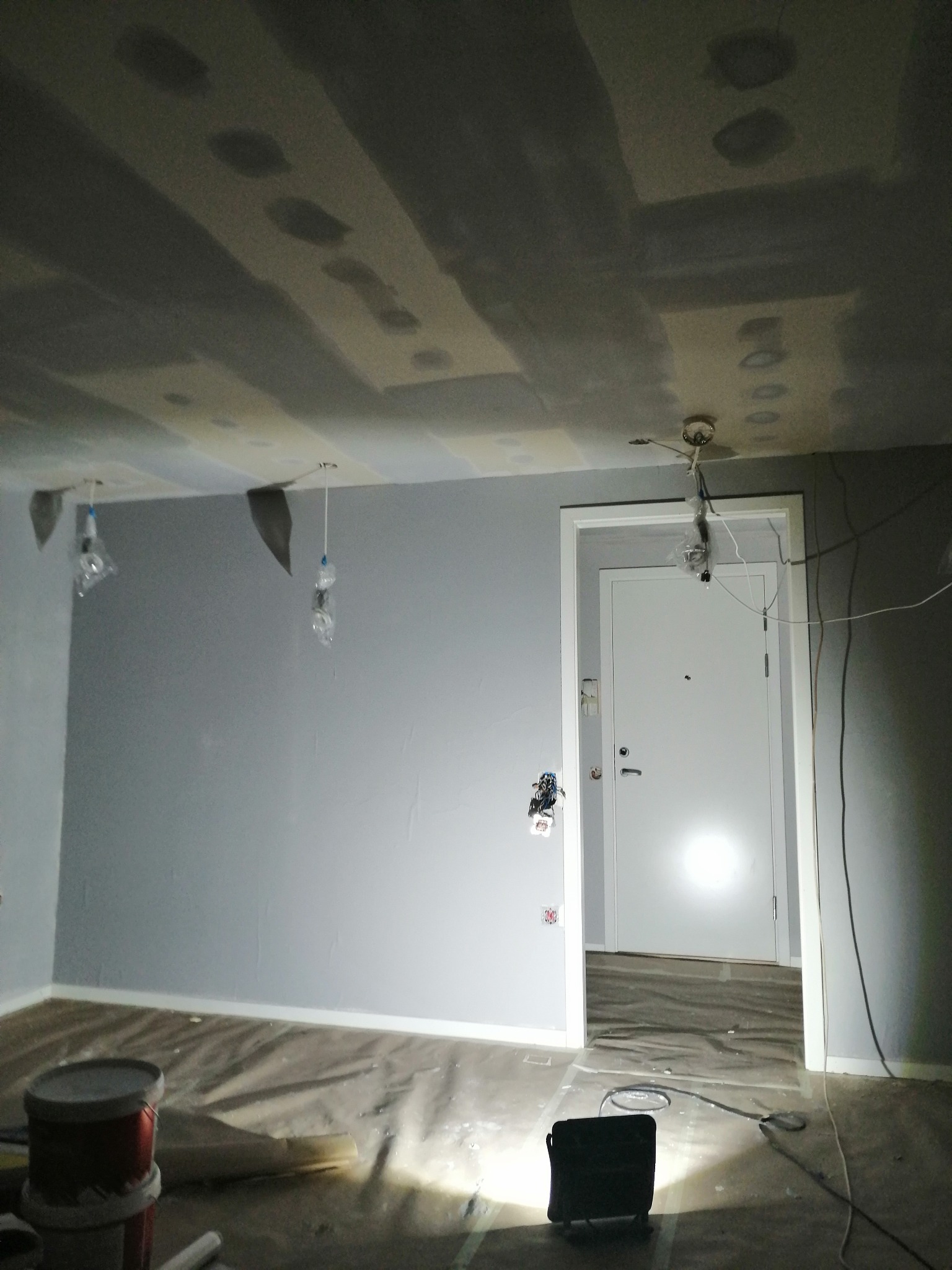 Pokój w trakcie remontu: szpachlowane i malowane ściany na szaro, zabezpieczona podłoga folią, widoczne puszki z farbą, przygotowane punkty oświetleniowe