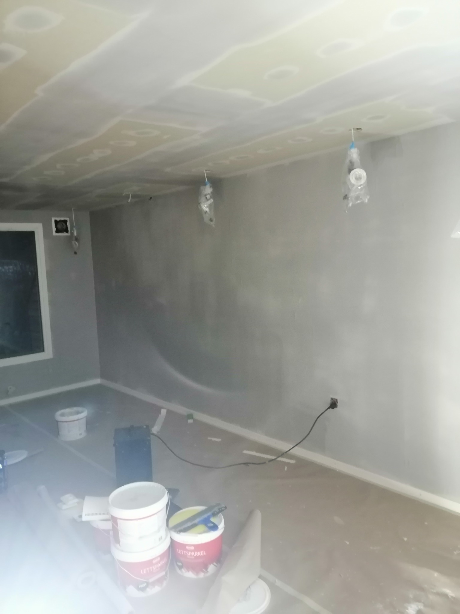Wnętrze pokoju w trakcie prac wykończeniowych, ściany pokryte świeżo nałożoną szpachlą, widoczne puszki z masą szpachlową i narzędzia malarskie, zabezpieczone folią oświetlenie sufitowe.