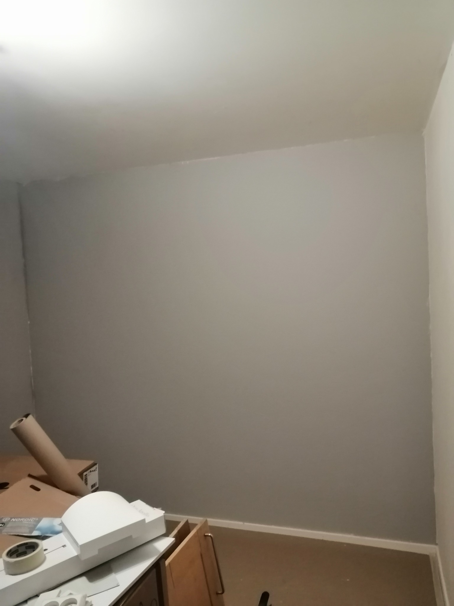 Świeżo pomalowana na szaro ściana w pokoju; widoczne kartony i akcesoria malarskie na podłodze, sugerujące prace wykończeniowe.