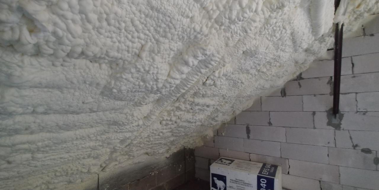 Poddasze z nierównomiernie nałożoną, grubą warstwą białej pianki izolacyjnej, widoczne fragmenty ściany z białego betonu komórkowego i kartonowe opakowanie.