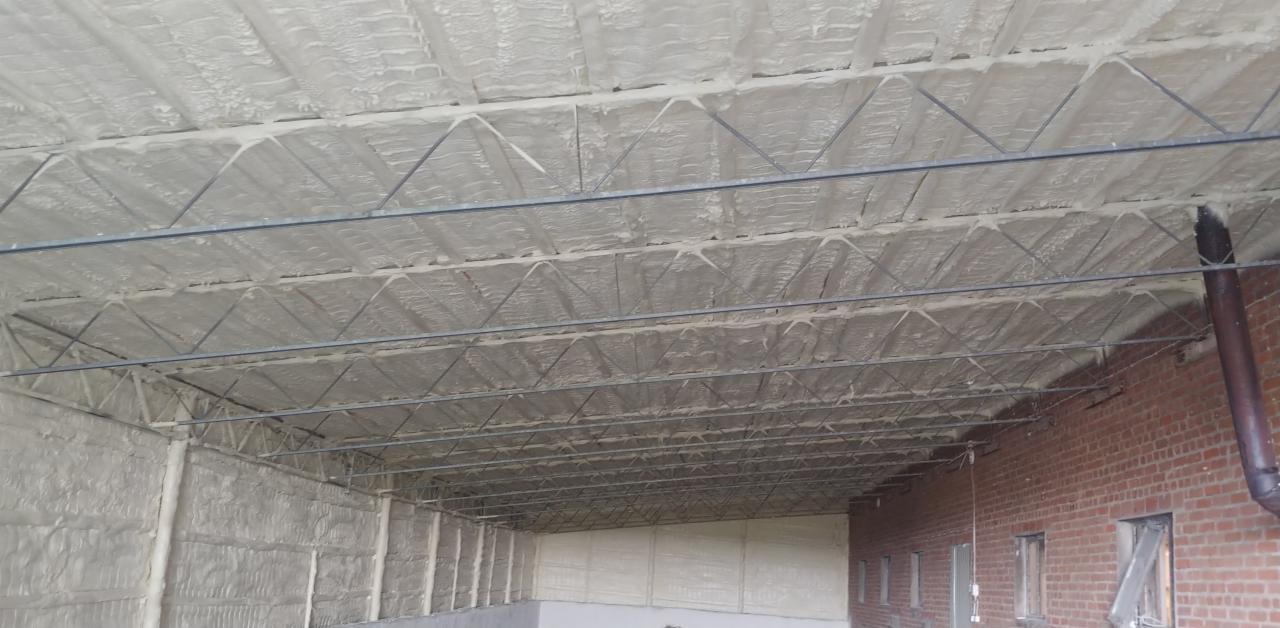 Poddasze przemysłowe po ociepleniu pianką PUR, widoczne metalowe elementy konstrukcyjne i fragment ściany z czerwonej cegły z rurą spustową.