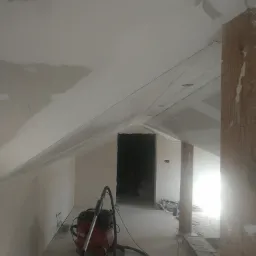 Poddasze w trakcie szpachlowania, widoczna szlifierka do gładzi na podłodze, sufit ze skośnymi ścianami częściowo pokryty gładzią, w tle otwór drzwiowy.