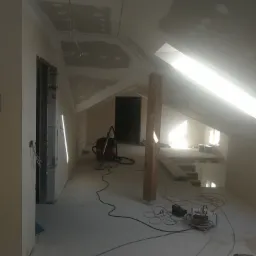 Poddasze w trakcie prac wykończeniowych, widoczne niedokończone gładzie na skosach, wystające kable, lampy budowlane, odkurzacz przemysłowy i drewniany słup konstrukcyjny.