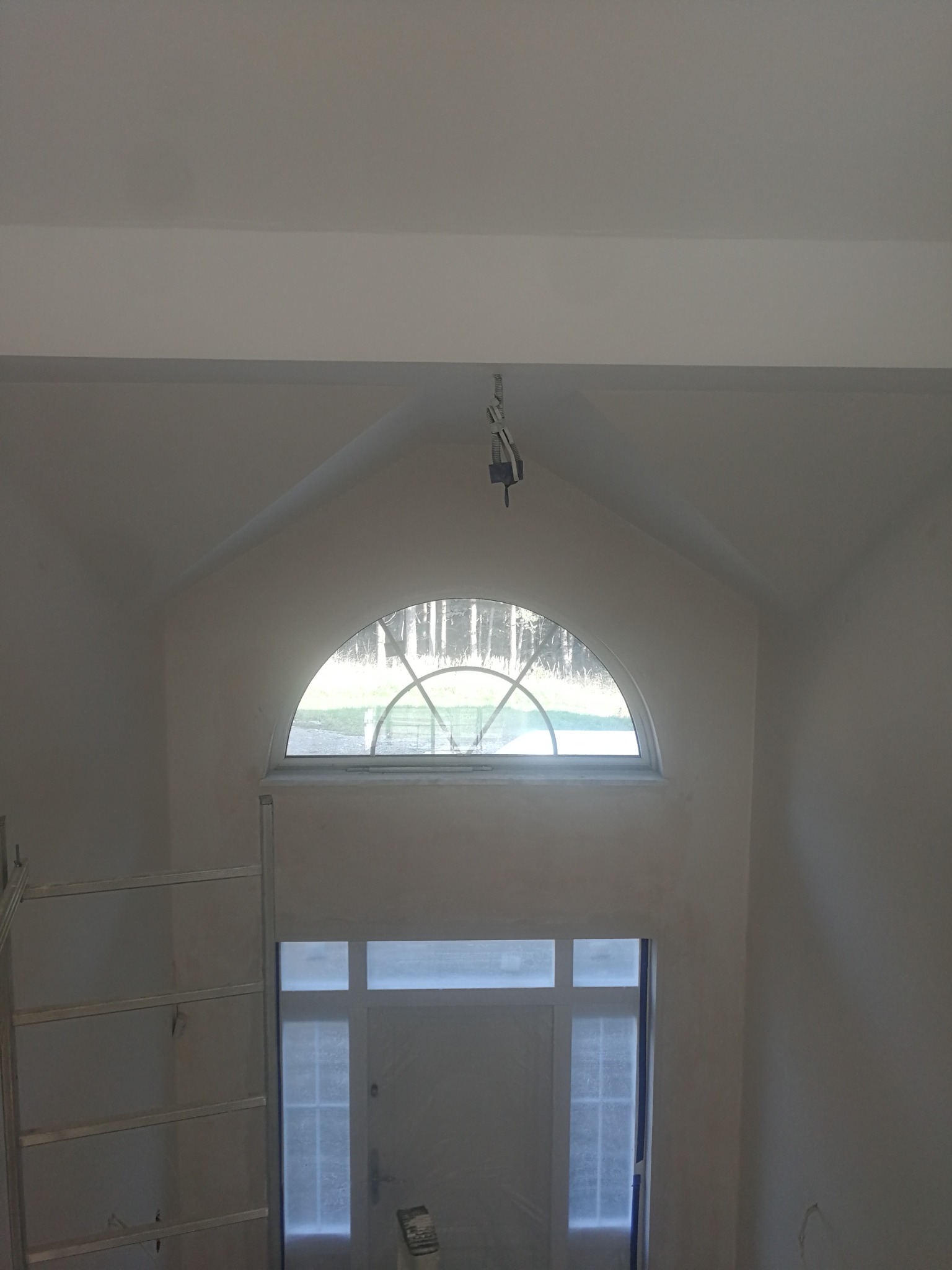 Wnętrze w trakcie malowania, widoczne świeżo zagruntowane ściany, okno półokrągłe z widokiem na zewnątrz, drabina aluminiowa i przewody oświetleniowe zwisające z sufitu.