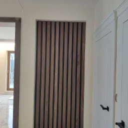 Nowoczesna aranżacja ściany z paneli dekoracyjnych z drewna w pionowe pasy, sąsiadująca z białymi drzwiami z czarnymi klamkami i fragmentem marmurowej podłogi.