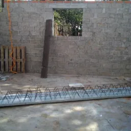 Wznoszona ściana z szarego bloczka z otworem okiennym, widoczne zbrojenie nadproża, paleta budowlana i pionowa deska oparta o ścianę.