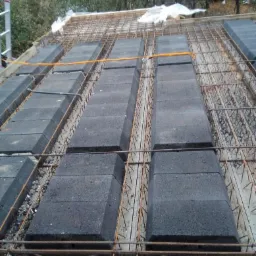 Strop Teriva w trakcie montażu, widoczne bloczki betonowe ułożone pomiędzy żebrami, zbrojenie siatką stalową i pomarańczowy przewód elektryczny, w tle zieleń drzew.