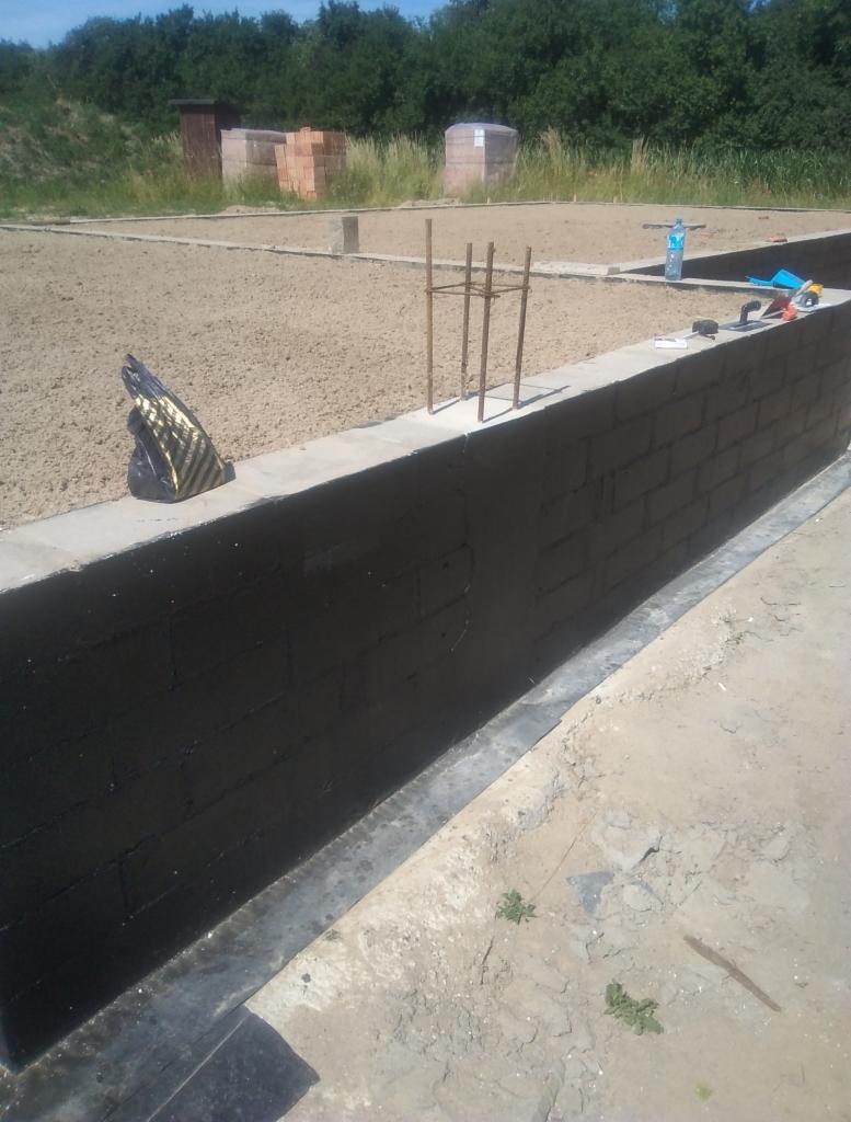 Zabezpieczone, czarną izolacją, murowane ściany fundamentowe z widocznym zbrojeniem, zasypane piaskiem w wykopie pod budowę.