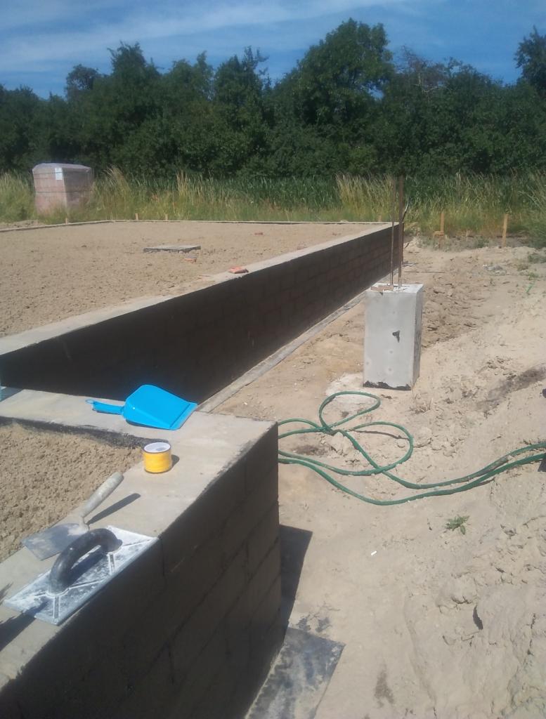 Wznoszone mury fundamentowe z cegły na placu budowy, widoczne narzędzia murarskie, niebieska szufelka i zielony wąż ogrodowy. W tle zarys lasu.