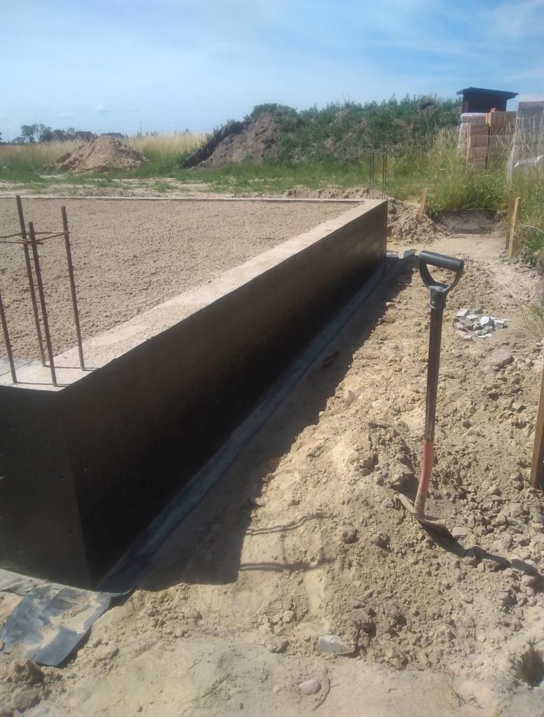 Wylany fundament z hydroizolacją, wystające pręty zbrojeniowe, łopata wbita w ziemię obok, widok na plac budowy.