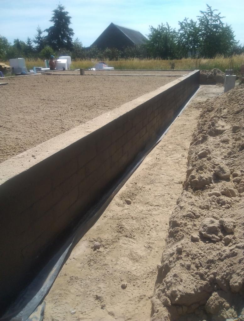 Wznoszenie fundamentów z bloczków betonowych, widoczna hydroizolacja, zasypana piaskiem przestrzeń wewnątrz fundamentu, w tle teren budowy z materiałami budowlanymi i budynkiem.