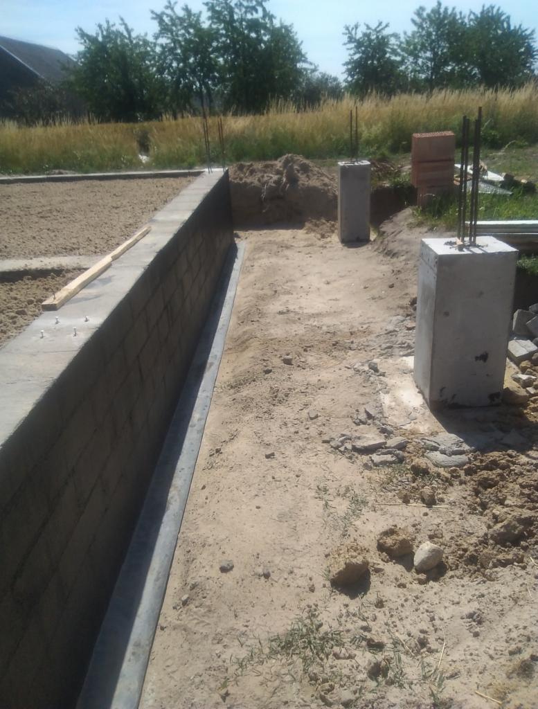 Wykop pod fundament, ściana z bloczków betonowych z izolacją pionową, betonowe słupki z wystającymi prętami zbrojeniowymi, piasek zasypowy, teren budowy.