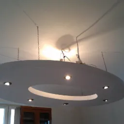 Okrągła, podwieszana konstrukcja z płyty karton gips z wbudowanymi punktami świetlnymi, zawieszona na łańcuchach, widoczne kable i instalacja elektryczna na suficie.