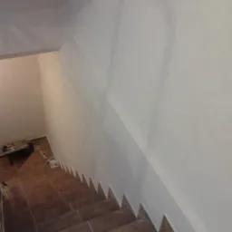 Klatka schodowa po malowaniu ścian na biało, widoczne brązowe płytki na schodach i narzędzia malarskie w rogu.