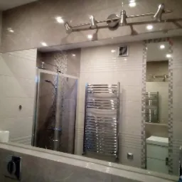 Beżowa łazienka z kabiną prysznicową, grzejnikiem drabinkowym i lustrem odbijającym fragment drugiej łazienki, widoczne dwie rolki papieru toaletowego.