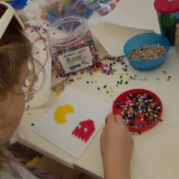 Dziewczynka tworzy obrazek Pac-Mana z kolorowych koralików na białej podkładce, otoczona pojemnikami z koralikami i narzędziami na jasnym stole.