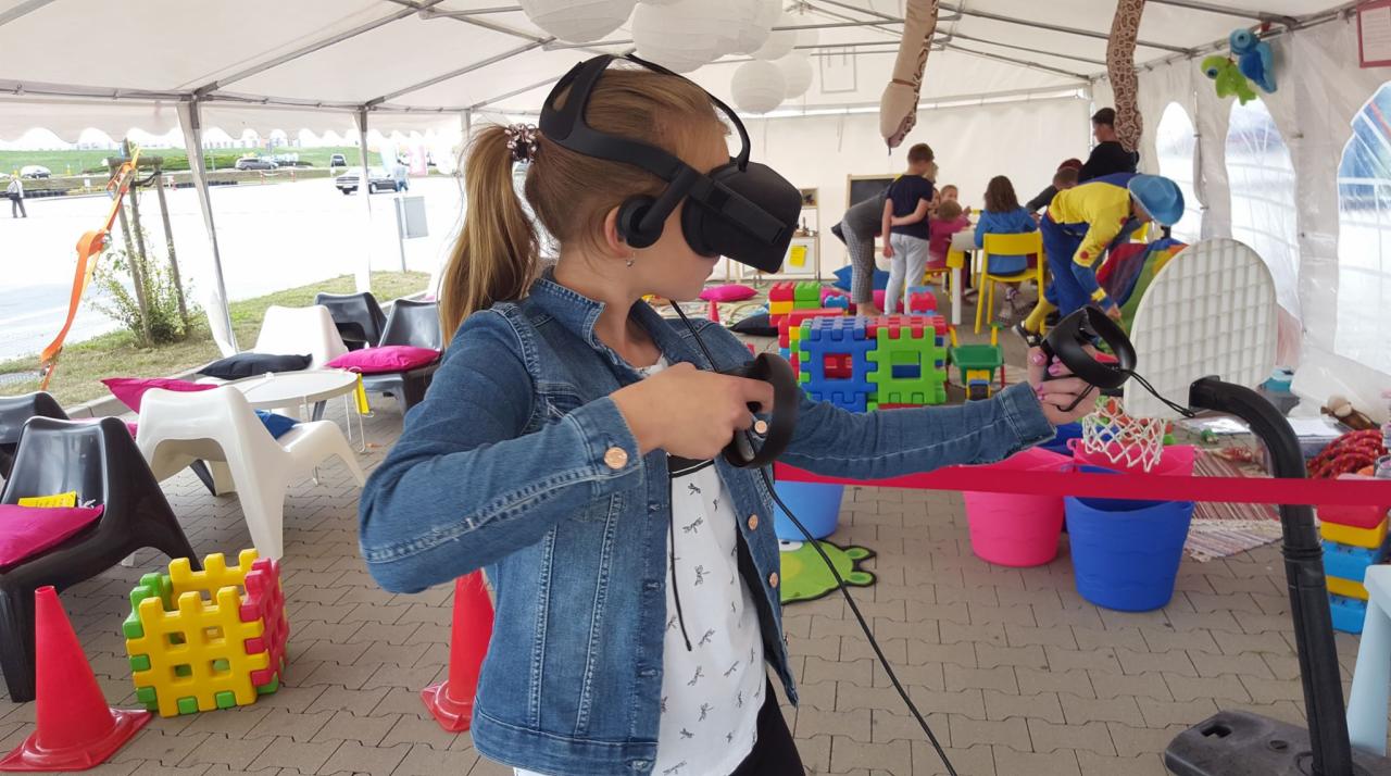 Dziewczynka w dżinsowej kurtce i goglach VR trzyma kontrolery w rękach, bawiąc się w wirtualnej rzeczywistości na tle kolorowych zabawek, dmuchanych dekoracji i innych dzieci w namiocie.
