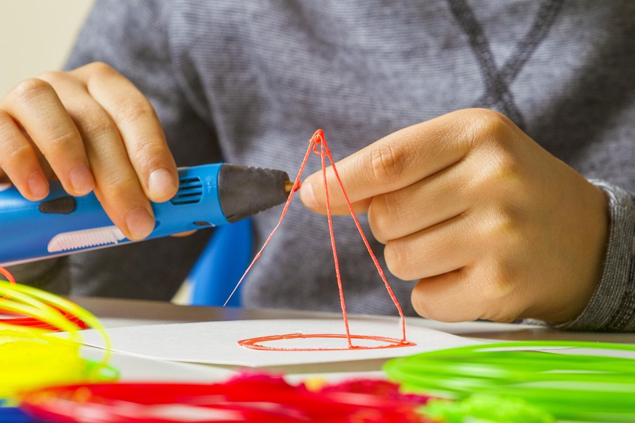 Ręce dziecka tworzą trójwymiarową konstrukcję piramidy z pomarańczowego plastiku za pomocą niebieskiego długopisu 3D, na białym papierze, z kolorowymi wkładami do długopisu w tle.