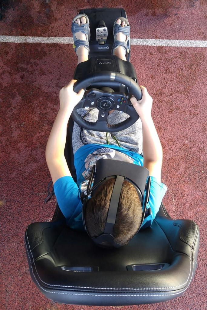 Chłopiec w sandałach siedzi na czarnym fotelu, trzyma kierownicę i ma na głowie gogle VR, symulator wyścigów na czerwonym tle.