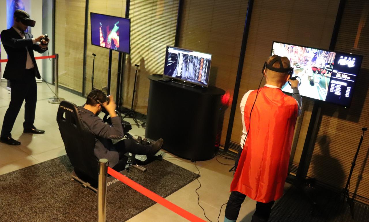 Trzech mężczyzn korzysta z zestawów VR: jeden w garniturze stoi, drugi siedzi w fotelu symulatora, trzeci w czerwonej pelerynie gra na stojąco przed ekranem.