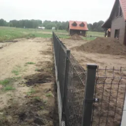 Nowo postawione, czarne ogrodzenie panelowe z widocznymi słupkami i siatką, na tle budynków w trakcie budowy i ziemi z wykopu.