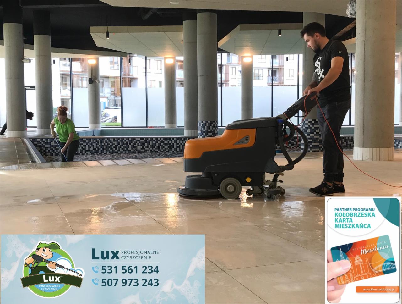 Pracownik obsługuje pomarańczowo-czarną maszynę do czyszczenia podłóg w przestronnym wnętrzu z kolumnami, podczas gdy druga osoba czyści krawędzie basenu. Podłoga jest mokra i błyszcząca.