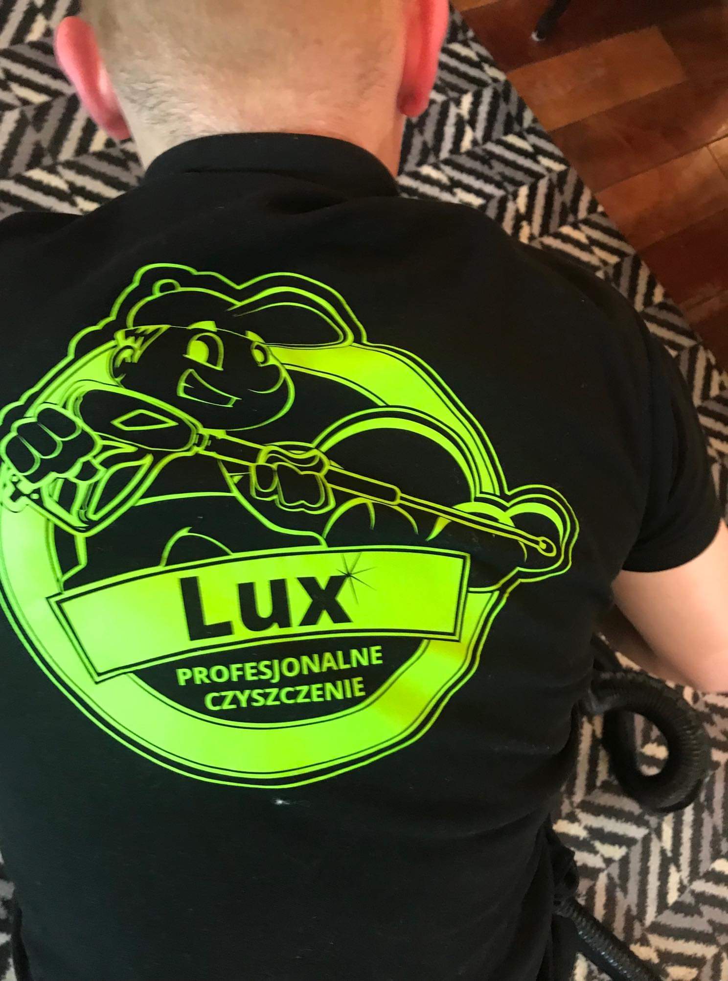 Czarna koszulka z neonowo-zielonym logo firmy sprzątającej 'Lux Profesjonalne Czyszczenie' z wizerunkiem królika z lancą ciśnieniową na tle dywanu.