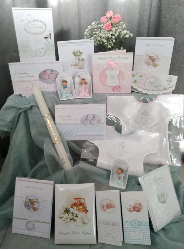 Komplet pamiątek Chrztu Świętego: kartki okolicznościowe, świeca z dekoracją, ubranko, bib i figurki aniołków, wszystko w pastelowych barwach na tle materiału.