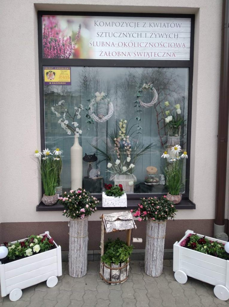 Wystawa kwiaciarni w Mińsku Mazowieckim z kompozycjami kwiatowymi w donicach, wazonach i drewnianych skrzynkach, widoczne dekoracje ślubne i żałobne.