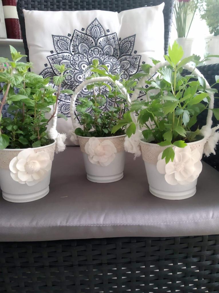 Trzy białe doniczki z roślinami, ozdobione koronką i kwiatami z materiału, ustawione na szarym siedzisku z poduszką z motywem mandali w tle.