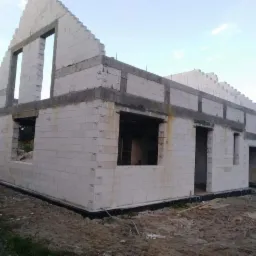 Niedokończony dom z białego pustaka z widocznymi otworami na okna i drzwi. Szary wieniec na ścianach. W tle zielone drzewa i błękitne niebo.