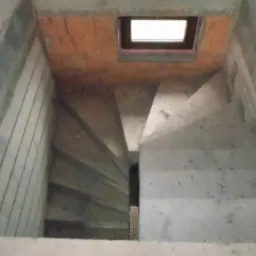 Wnętrze piwnicy w trakcie budowy z widocznymi betonowymi schodami i murem z pustaków, oświetlone przez niewielkie okno w suficie.